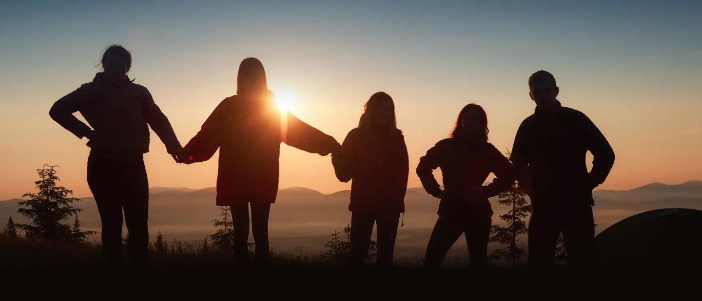 Fem mennesker står på rad og række udendørs ved solnedgang og holder hinanden i hånden. Deres silhuetter ses mod himlen med bakker i baggrunden.