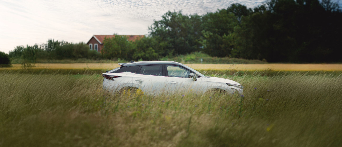 En hvid OMODA EV 5 parkeret på en græsmark med træer og et hus i baggrunden under en delvis overskyet himmel.